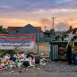 Indonesia bebas sampah 2029: warga memilah sampah plastik dan kertas di lingkungan perumahan dengan latar tumpukan sampah dan banner kampanye kesiapan Indonesia bersih 2029